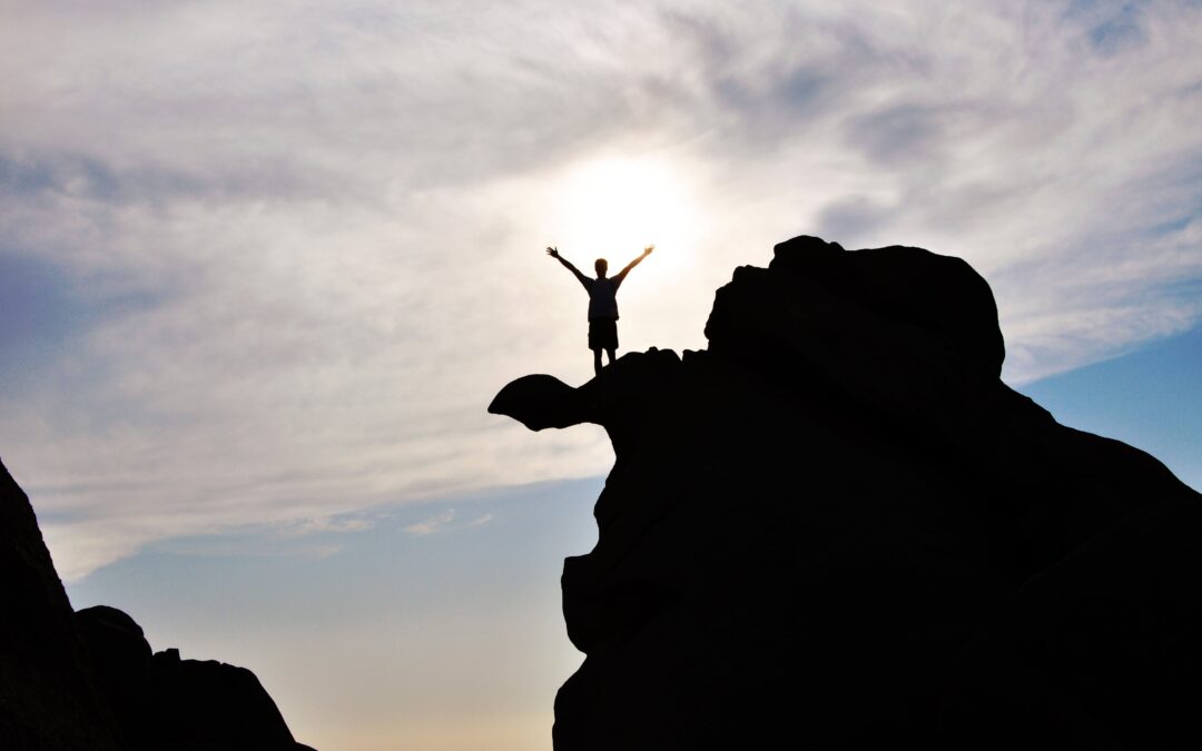 A person is standing on top of a rock with their arms over their head in triumph. They are looking out at the sky.